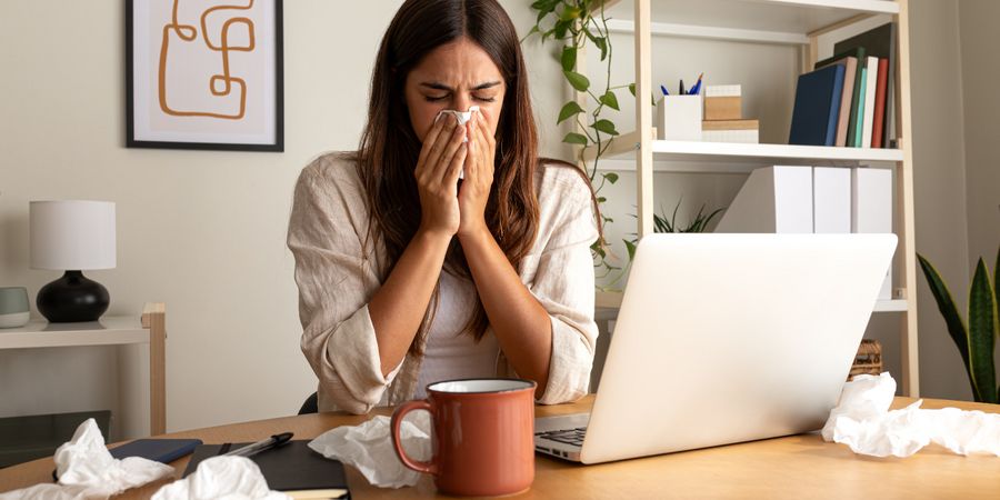 Bild: Frau arbeitet im Home Office, niest und hat einige Taschentücher auf dem Schreibtisch liegen. Schimmel vorbeugen ist wichtig, da er Allergien auslösen kann mit unspezifischen Symptomen.