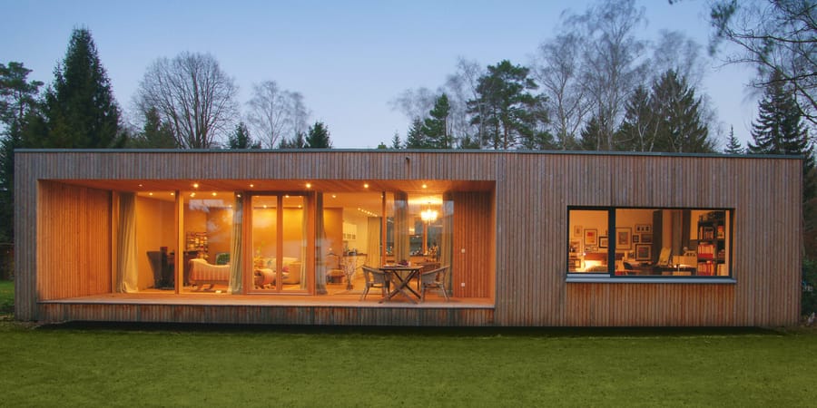 Nachhaltiger Architekten Bungalow mit Holzverschalung und großen Fenstern zur Gartenseite hin. Der Grundriss ist rechteckig. 