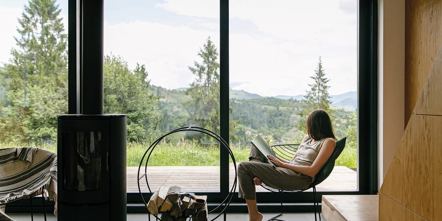 Frau liest ein Buch in einem Sessel vor einem bodentiefen Fenster und blickt in einen Wald.