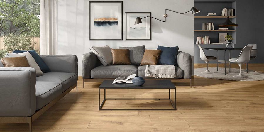 Wohnzimmer mit hellen Wänden und grauem Sofa mit Fliesen in Holzoptik am Boden. 