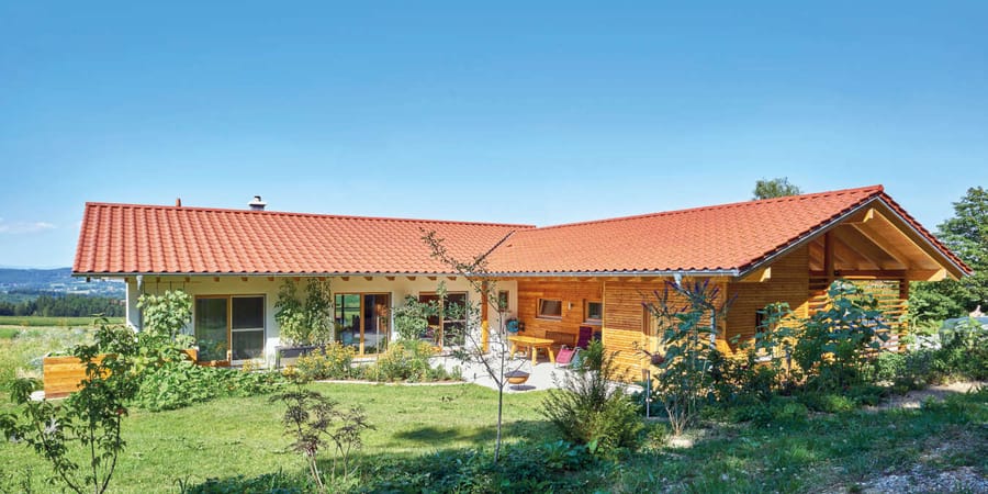 Bungalow in L-Form mit rotem Satteldach und Holzfassade. Die Grundfläche und der Grundriss des  Wohnbereichs sind rechteckig. Die L-Form entsteht durch eine angegliederte Doppelgarage